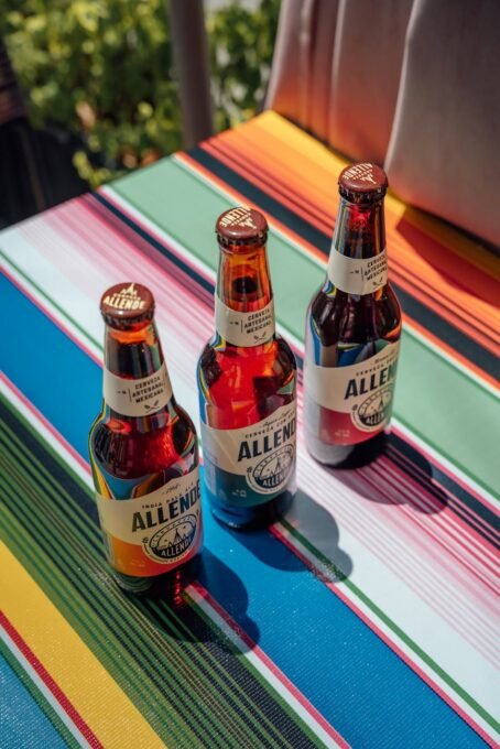 Three Allende beer bottles on a vibrant Mexican tablecloth in sunlight.